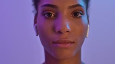 Kulaklıklı güzel Afrikalı bir kadının mor neon arka planda yalnız kalmaktan korkmasını yakından izleyin.
