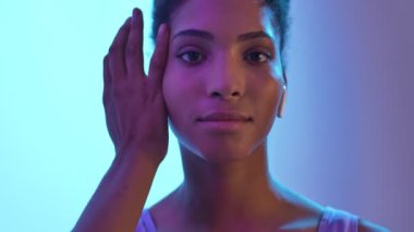 Kulaklıklı, mutlu bir Afrikalı kadının yüzüne dokunuşunu ve bej renkli neon arka planda izole edilmiş kameraya bakışını kapatın.