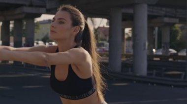 Spor sutyenli, sakin genç bir kadın, şehir köprüsünün yakınındaki eğitimden önce sırtını esnetiyor.