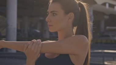 Spor sutyenli hoş sakin genç bir kadın antrenmandan önce kollarını uzatıyor sonra da şehrin yakınındaki köprünün dışındaki kameraya bakıyor.