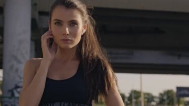 Ciddi şekilde konsantre olmuş genç bir kadın, şehir köprüsünün yakınındaki eğitim sırasında kulaklıklarına dokunuyor.