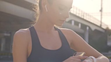 Spor kıyafetli, kulaklık takan bir kadın şehirde antrenman yaparken spor takip cihazını kullanıyor.