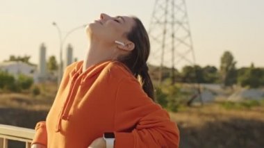 Güzel bir kadın, köprüdeki korkuluklara yaslanıp güneşli bir sabahta açık havada müzik dinlerken dinleniyor.