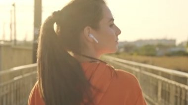 Kablosuz kulaklıklı sakin ve rahat genç bir kadının arka görüntüsü güneşli bir sabahta köprüde yürüyor.