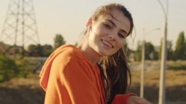Spor kıyafetleri içinde gülümseyen genç bir kadın sabah köprüde güneşe doğru dönerken korkulukların yanında duruyor.