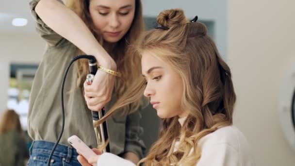 Professional Hairdresser Doing Curls Using Iron While Client Using Smartphone — Stock Video
