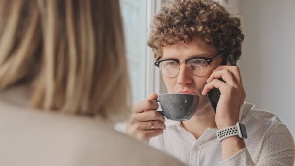一个戴着眼镜的迷人的卷发男人一边坐在咖啡店一边打电话 一边和同事一起在屋里喝咖啡 — 图库视频影像