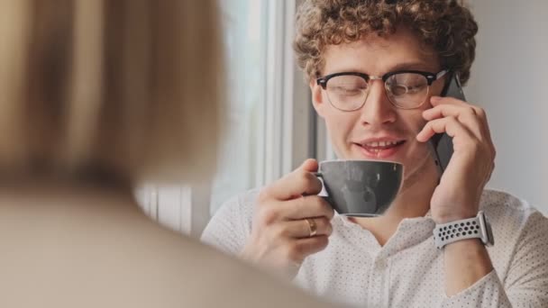 一个戴着眼镜的积极卷曲的男人正在和他的同事在咖啡馆里通电话 在屋里喝咖啡 — 图库视频影像