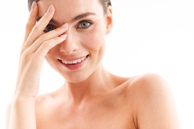 Çekici, gülümseyen, üstsüz, esmer bir kadının güzel portresi beyaz arka planda izole bir şekilde duruyor, yüzüne dokunuyor, kameraya bakıyor.