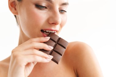 Çekici, gülümseyen, üstsüz, esmer bir kadının portresi. Beyaz arka planda tek başına duruyor. Çikolata tabağını ısırıyor.