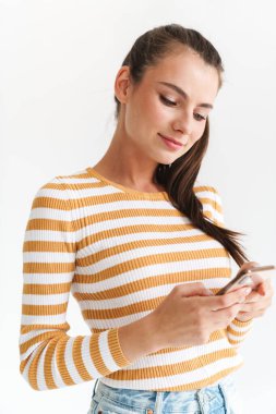 Gülümseyen, günlük kıyafetler giyen, beyaz arka planda tek başına dururken cep telefonu kullanan genç bir kız.