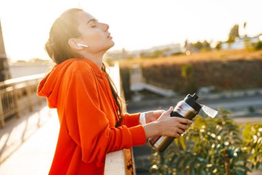 Dışarıda kulaklıklı, su dolu bir şişeyle müzik dinleyen çok güzel bir spor fitness kadınının fotoğrafı..