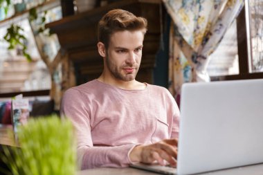 Kafede otururken dizüstü bilgisayarda çalışan yakışıklı dalgın genç adam.