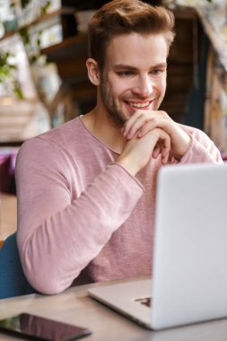 Kafede otururken dizüstü bilgisayarda çalışan yakışıklı, gülümseyen genç adam.