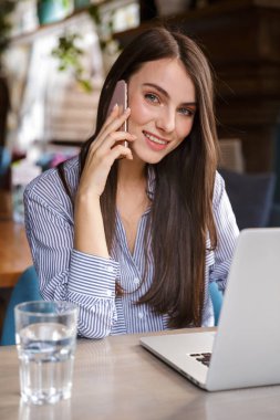 Çekici, gülümseyen, esmer bir kadın, kafede oturmuş cep telefonuyla konuşurken dizüstü bilgisayarla çalışıyor.