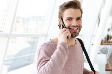 Çantalı genç sakallı bir adamın parlak pencerenin önünde akıllı bir telefonla konuşurken görüntüsü.