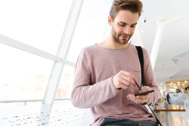 Elinde çanta olan genç sakallı bir adamın parlak pencerenin önünde dikilirken akıllı telefonu tutuşu.