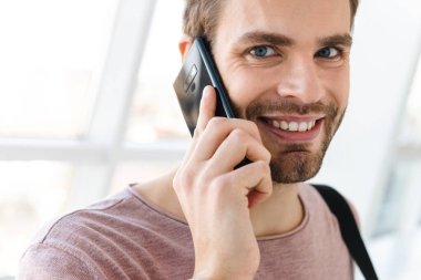 Çantalı genç sakallı bir adamın parlak pencerenin önünde akıllı bir telefonla konuşurken görüntüsü.