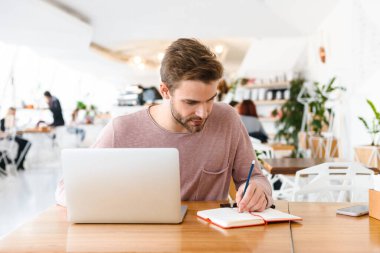 Kafede otururken dizüstü bilgisayarda çalışan sakallı bir genç resmi.
