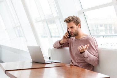 Kafede pencere kenarında otururken akıllı telefon ve dizüstü bilgisayar kullanan sakallı bir genç resmi.