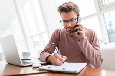 Cep telefonuyla konuşan ve kafenin içinde kağıt belgelerle çalışan genç sakallı bir adam.