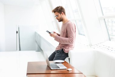 Gözlük takan, akıllı telefon kullanan ve kapalı kafede dizüstü bilgisayarda çalışan genç sakallı bir adam.