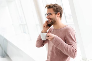 Gözlük takan genç sakallı bir adamın parlak pencerenin önünde dikilirken akıllı telefondan konuşması.