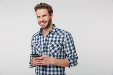 Ekose gömlek giyen, gülümseyen ve beyaz arka planda izole edilmiş cep telefonu kullanan mutlu genç adamın portresi.