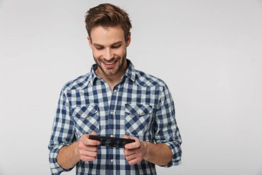 Ekose gömlek giyen, gülümseyen ve cep telefonuyla oyun oynayan mutlu bir gencin portresi beyaz arka planda izole edilmiş.