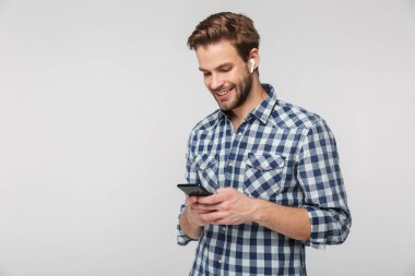 Ekose gömlek giyen mutlu bir gencin portresi. Beyaz arka planda izole edilmiş cep telefonu ve kulaklık kullanıyor.