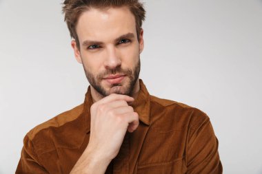 Tıraşsız, kahverengi gömlekli bir gencin portresi beyaz arka planda izole edilmiş kameraya bakıyor ve poz veriyor.