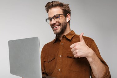 Gözlüklü neşeli bir gencin portresi. Beyaz arka planda dizüstü bilgisayar ve kulaklık kullanırken başparmağını gösteriyor.