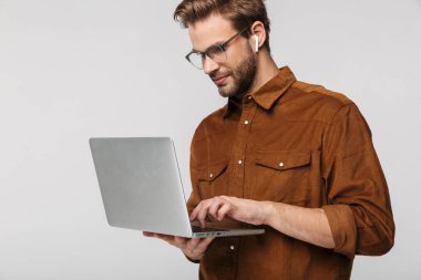 Gözlüklü yakışıklı adamın portresi beyaz arka planda izole edilmiş dizüstü bilgisayar ve kulaklık kullanıyor.