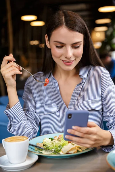 Bela Jovem Sorridente Usando Telefone Celular Enquanto Almoça Café Dentro — Fotografia de Stock