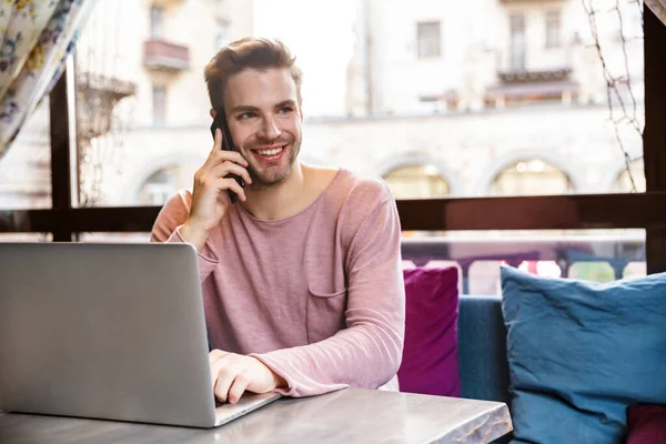 Przystojny Uśmiechnięty Młodzieniec Pracujący Laptopie Siedząc Kawiarni Rozmawiając Przez Telefon — Zdjęcie stockowe