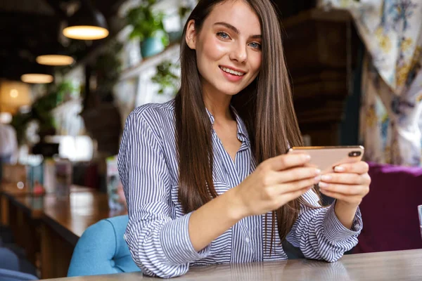 Linda Sorridente Jovem Morena Segurando Telefone Celular Enquanto Sentado Café — Fotografia de Stock