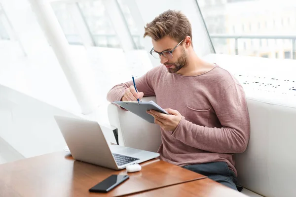 屋内カフェで仕事をしながらノートパソコンを使ってクリップボードを保持している若い髭の男のイメージ — ストック写真