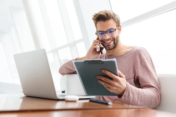 若い髭の男が携帯電話で話をし 屋内カフェで働いている間にクリップボードを保持するイメージ — ストック写真