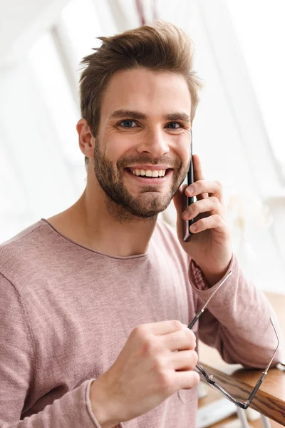 屋内で明るい窓の上に立っている間に眼鏡をかけ 携帯電話で話している若い髭の男のイメージ — ストック写真