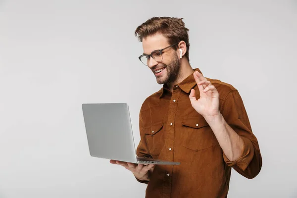 Portrait of cheerful young man in eyeglasses waving hand while using laptop and earphone isolated over white background