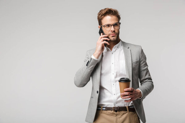 Portrait of serious handsome man in eyeglasses talking on cellphone and holding paper cup isolated over white background