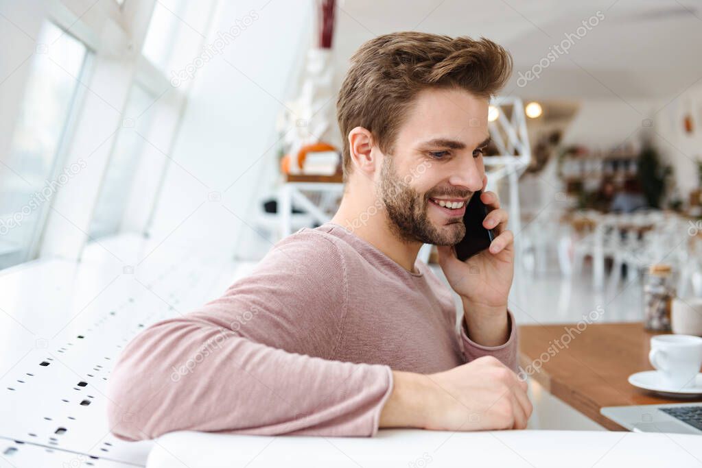 Image of young bearded man talking on smartphone while sitting on sofa by window in cafe indoors