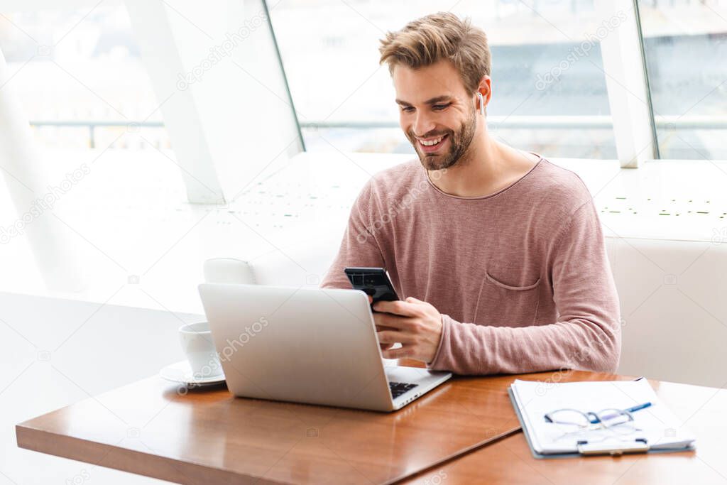 Image of young bearded man wearing earbuds working on laptop computer by window in cafe indoors
