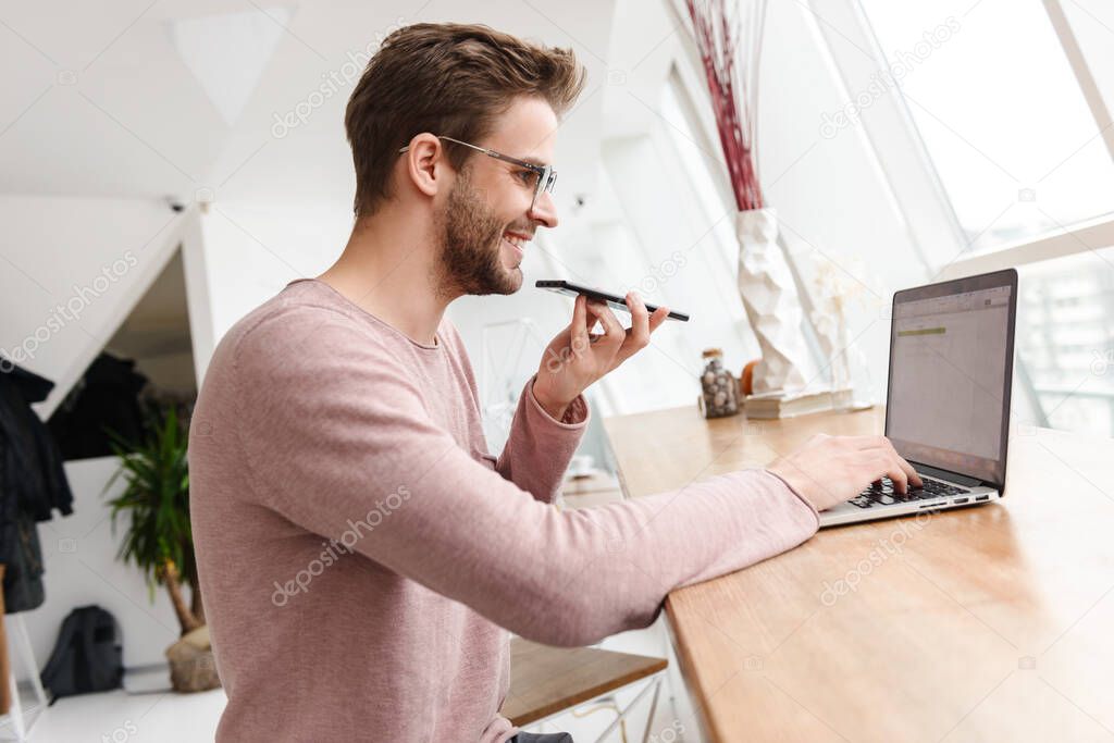 Image of young bearded man wearing eyeglasses working on laptop and using cellphone in cafe indoors