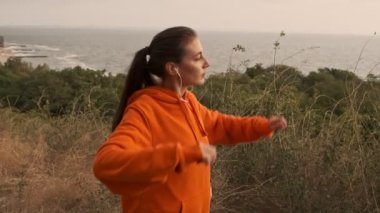 Tasasız, güzel, kulaklıklı, kollarıyla ısınan ve deniz kıyısına bakan atletik bir kadın.