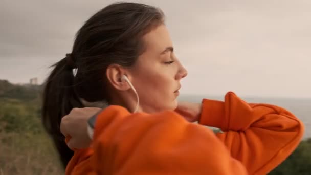 彼女のフードを修正し 海岸で実行する準備のイヤホンでかなり運動能力の女性 — ストック動画
