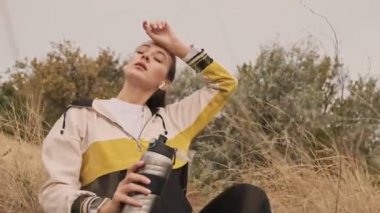 Kulaklıklı, yorgun ve atletik bir kadın antrenmandan sonra deniz kıyısında şişe ile otururken dinleniyor.