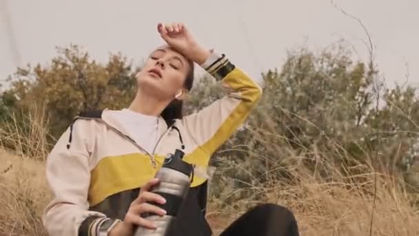 Zmęczona Dość Wysportowana Kobieta Słuchawkach Relaksująca Się Treningu Siedząc Butelką — Wideo stockowe