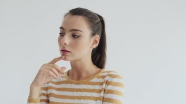 Genç ve güzel bir kadın stüdyoda beyaz arka planda tek başına dururken bir şeyler düşünüyor.