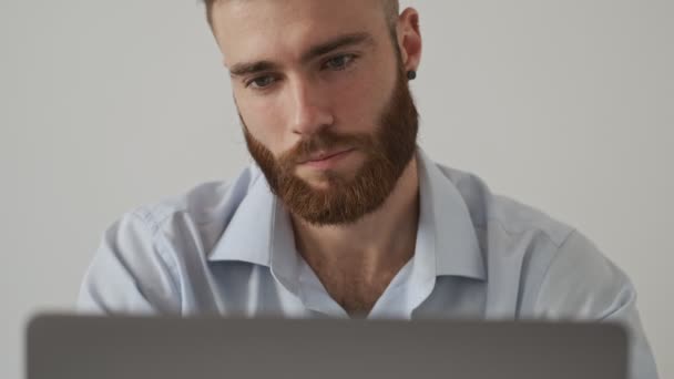 一位头戴衬衫 泰然自若的年轻大胡子男子正坐在白墙边的桌子旁 用笔记本电脑 — 图库视频影像